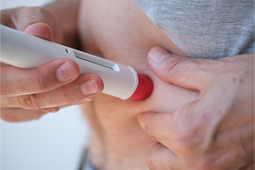 Close-up of a person pressing a small handheld medical device with a red tip against their abdomen, suggesting a needle-based injection system being applied to the skin.