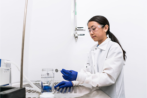 A woman in a white lab coat and blue gloves works at a laboratory bench, holding a small circular sample with tweezers over a perforated metal work surface. Nearby are a glass jar with liquid, lab equipment, and tubing against a clean, light-colored background.