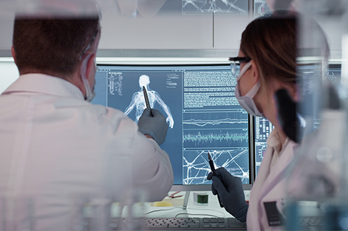Two people in a laboratory, wearing lab coats, face masks, and gloves, review medical imaging on a computer monitor. One person points to a full-body scan displayed on the screen while both hold pens, with additional charts and waveform data visible on the display.